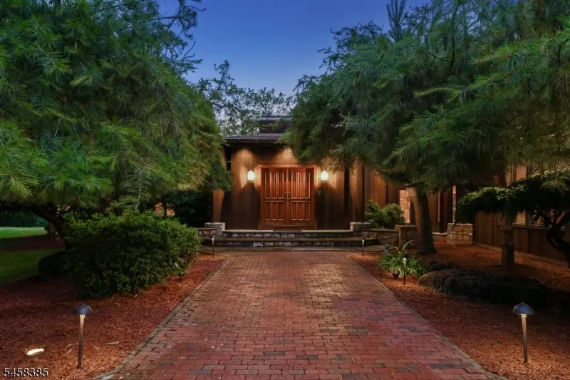 Twilight arrival at 51 Hoagland Road — a brick paver walkway lined with landscape lights leading to warmly lit wood double doors framed by mature pines